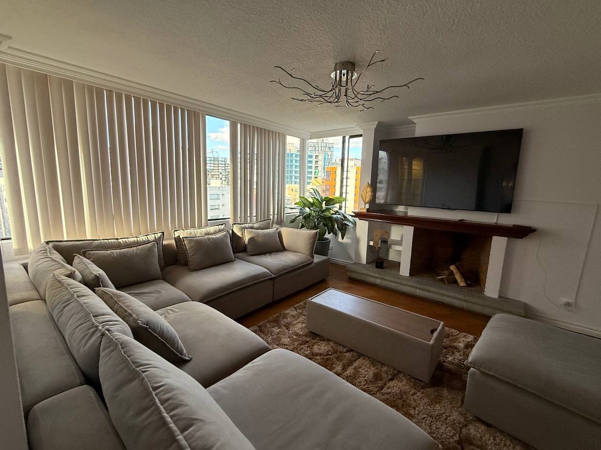A spacious living area is highlighted by a large, L-shaped sectional sofa, arranged comfortably around a central coffee table. Natural light pours in through expansive windows adorned with vertical blinds, offering views of the city. A flat-screen TV is mounted above a fireplace, complemented by decorative plants.