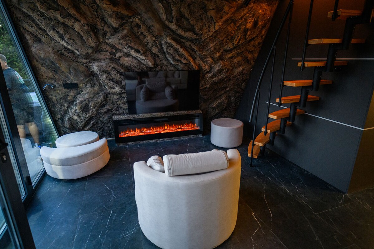 A modern living area showcases an elegant stone wall and a sleek television mounted above a modern electric fireplace. Two plush circular seats and a curved sofa are arranged to create a cozy conversation space. A staircase leads to the upper level, enhancing the sense of vertical space.