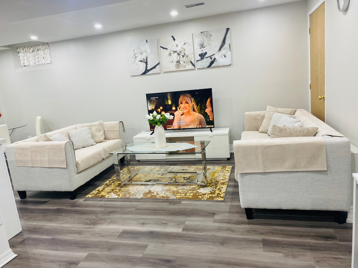 A modern living area is showcased with two matching light-colored sofas, positioned across from a low, rectangular coffee table. A decorative area rug in subtle hues adds warmth to the space. Wall art featuring floral designs adorns the walls, complemented by a flat-screen television.