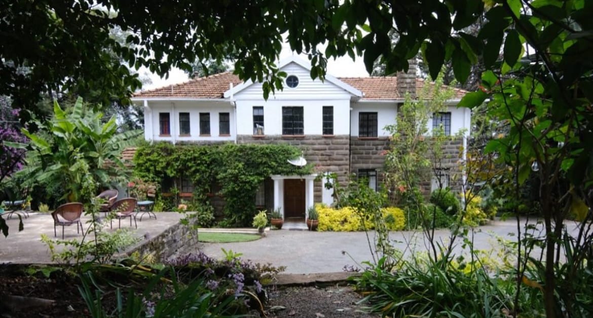 The exterior of Bustani Manor is captured amidst lush greenery, showcasing a two-story home with white walls and brown stone detailing. A garden blooms with vibrant flowers, while outdoor seating areas invite relaxation under the shade of surrounding trees.