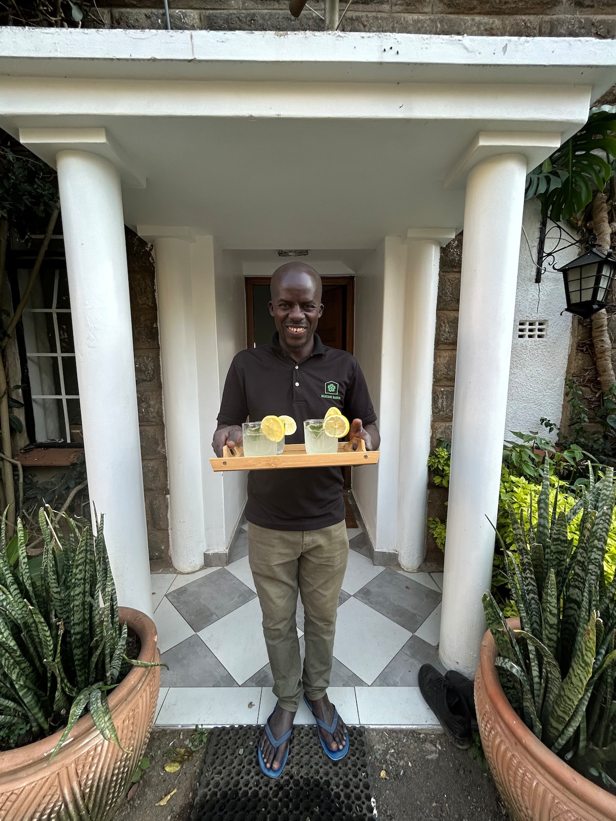 A server stands at the entrance of Bustani Manor, holding a wooden tray with two refreshing drinks garnished with lemon slices. The setting features classic white pillars and green plant arrangements on either side, enhancing the welcoming ambiance of the doorway.