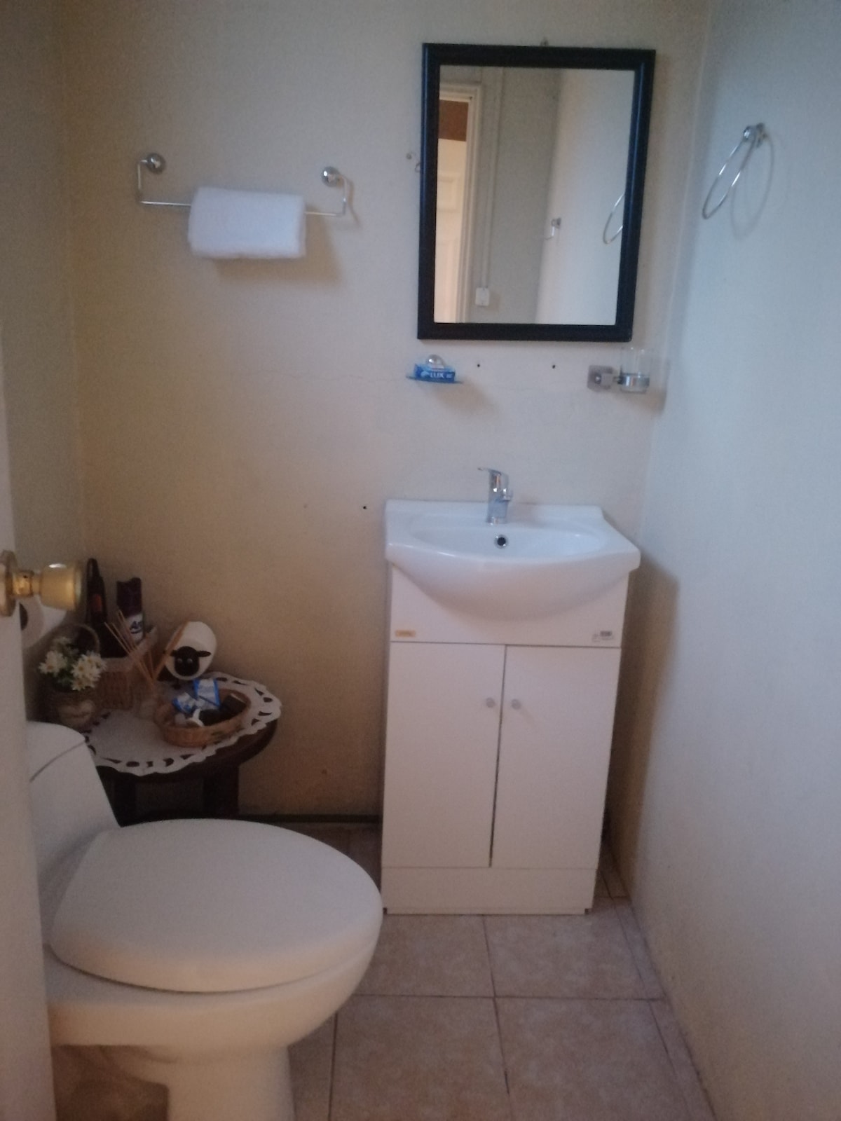 An organized bathroom is displayed featuring a compact sink with a mirror above it. A small countertop with decorative items complements the white toilet. Tiled flooring contributes to the neat appearance of the space, while hand towels are neatly hung on wall fixtures.