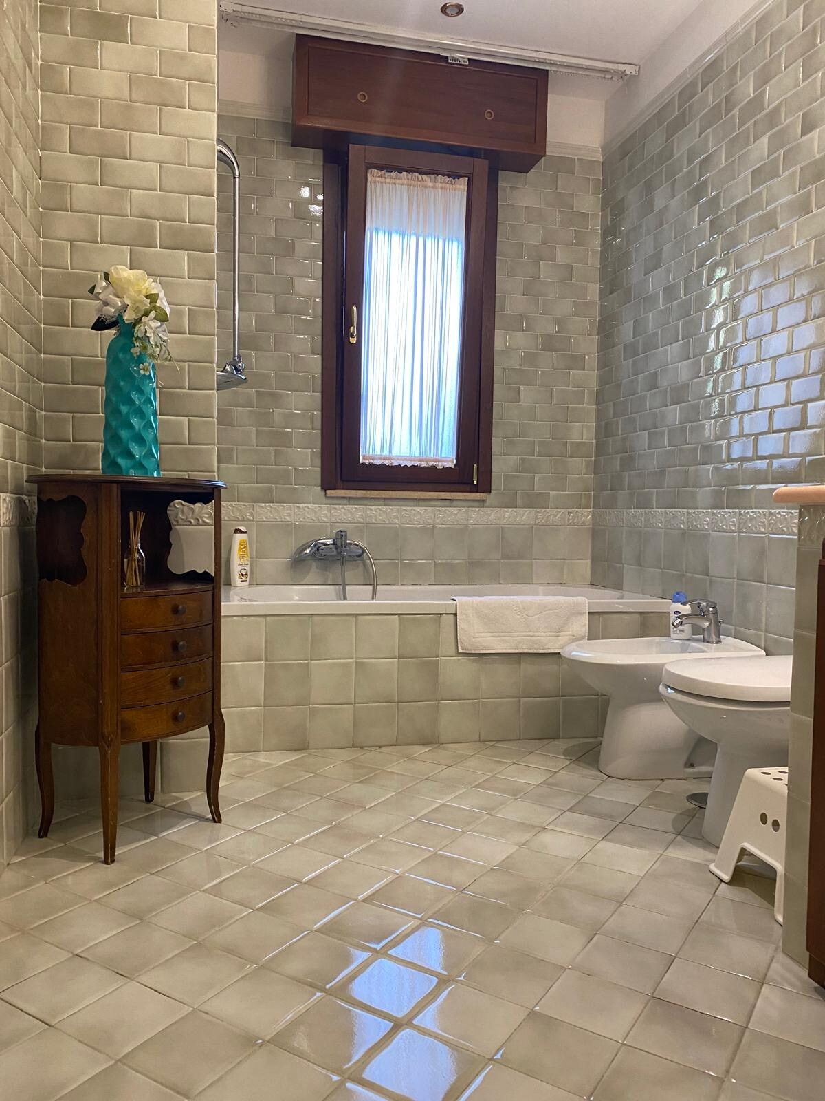The bathroom features light gray tiled walls and floor, allowing ample light to reflect across the space. A bathtub is positioned alongside a decorative wooden cabinet. A sink and toilet complete the layout, with a window providing natural light and a floral arrangement adding a touch of color.