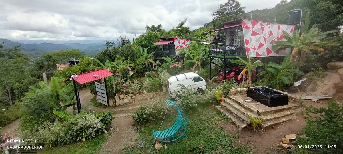 An outdoor view of the property shows elevated cabins surrounded by lush greenery and banana trees. A multi-level stone pathway leads to a shared hot tub. Colorful seating and a white vehicle are visible, with mountains in the background under a cloudy sky.