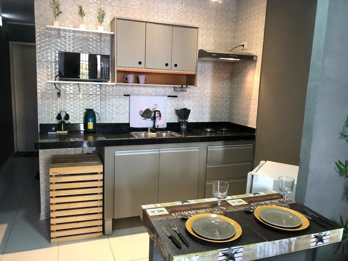 A modern kitchenette features a sleek design with neutral cabinetry and a dark granite countertop. There is a stainless steel sink, a stove with an overhead extractor, and thoughtfully arranged cooking utensils. A rustic dining table is set with two plates and glassware, creating a cozy dining area.
