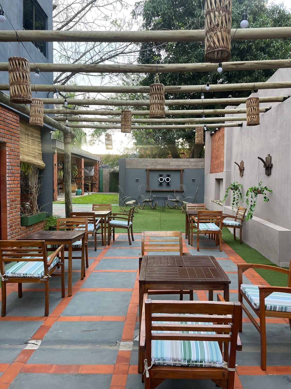 An outdoor seating area is furnished with wooden chairs and tables, arranged on a patterned floor. The overhead beams feature hanging lanterns and greenery, while a lush garden space is visible in the background, enhancing the inviting setting.