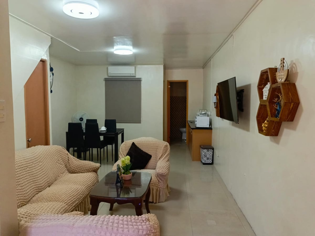 A living area is presented with a neutral color palette, featuring two cozy sofas and a wooden coffee table. A dining area with a dark table and black chairs is visible in the background, along with a cabinet holding various items. A wall-mounted TV is also included.