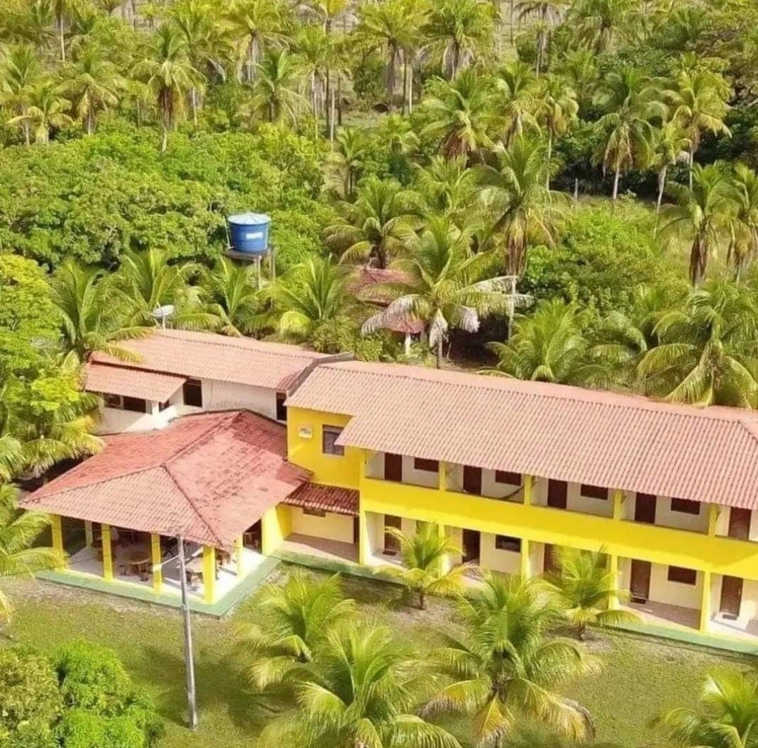 An aerial view showcases a rustic yellow building surrounded by lush greenery, featuring a terracotta roof. Palm trees and other vegetation fill the landscape, while a water tower stands in the background, highlighting the natural setting and expansive grounds.