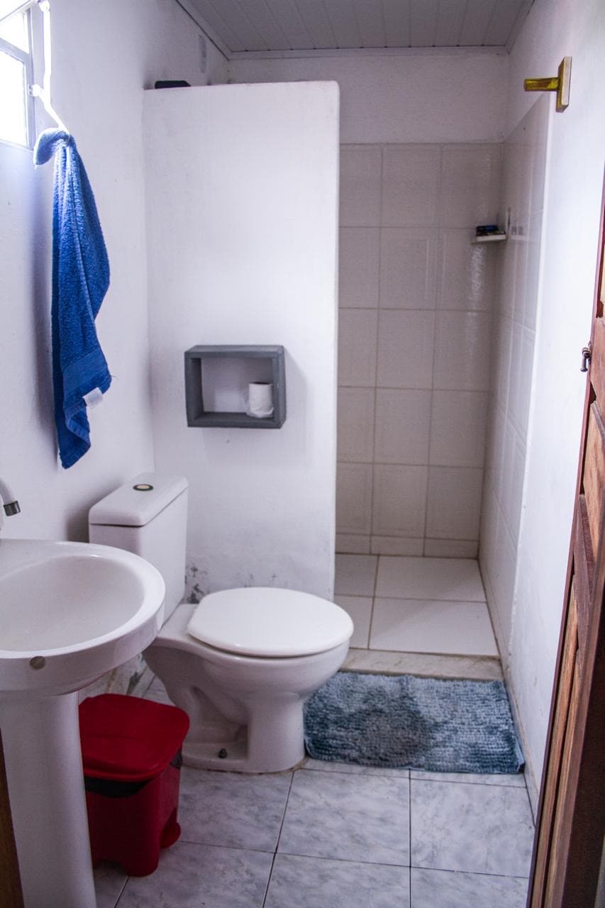 A clean and functional bathroom features a white toilet and a small pedestal sink. A shower is positioned in the corner, surrounded by tiled walls. A blue towel hangs on the wall, while a red bin and a gray rug add subtle touches to the space.