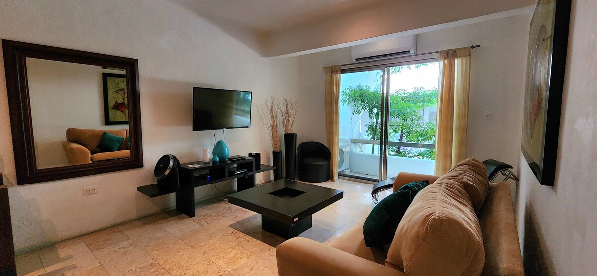 The living area features a spacious beige sofa complemented by colorful cushions. A large mirror reflects the room's layout, where a flat-screen television is positioned on a black media unit. Sliding glass doors lead to a balcony, allowing natural light to fill the space.