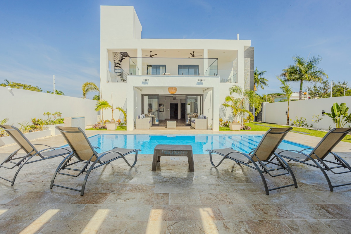A spacious outdoor area is highlighted by a crystal-clear pool, surrounded by six loungers positioned for relaxation. The villa's modern architecture features a second-floor balcony, offering a view of lush greenery and palm trees, contributing to a tranquil atmosphere.