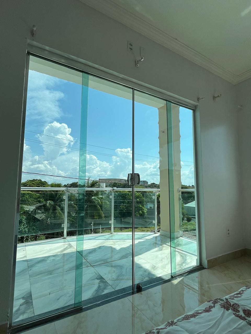 A large glass sliding door opens to a spacious balcony, allowing natural light to fill the room. The view beyond features a clear blue sky with scattered clouds, framed by the modern glass railings of the balcony.