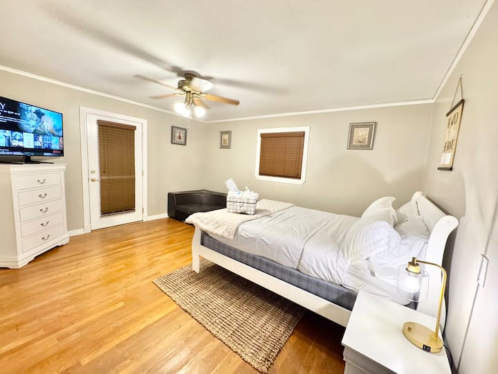 This inviting bedroom combines simplicity and comfort with bright, airy touches. The white bed frame and crisp linens create a clean, modern feel, complemented by soft golden lighting and warm hardwood floors. 