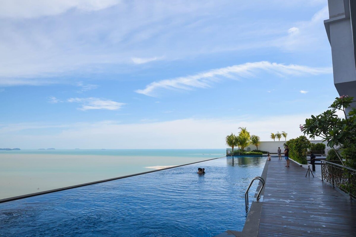 An infinity pool extends towards the horizon, blending with the serene blue waters of the sea. Clear skies above create a peaceful ambiance, while palm trees line the edge, adding greenery to the stunning view available from the high-rise setting.