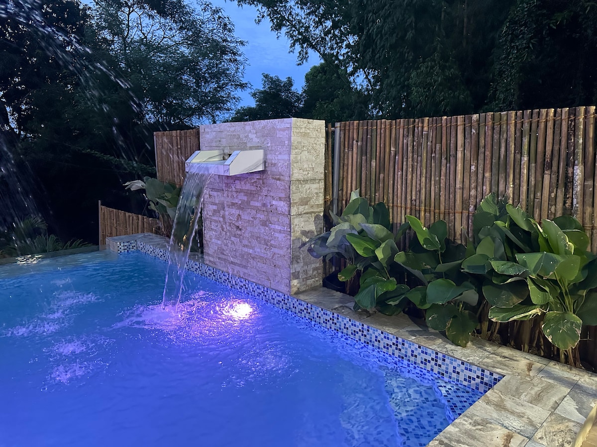 The infinity swimming pool is adorned with a soothing fountain feature, surrounded by lush greenery and a bamboo fence. Evening lighting casts a gentle glow on the water, enhancing the tranquil ambiance of the space.