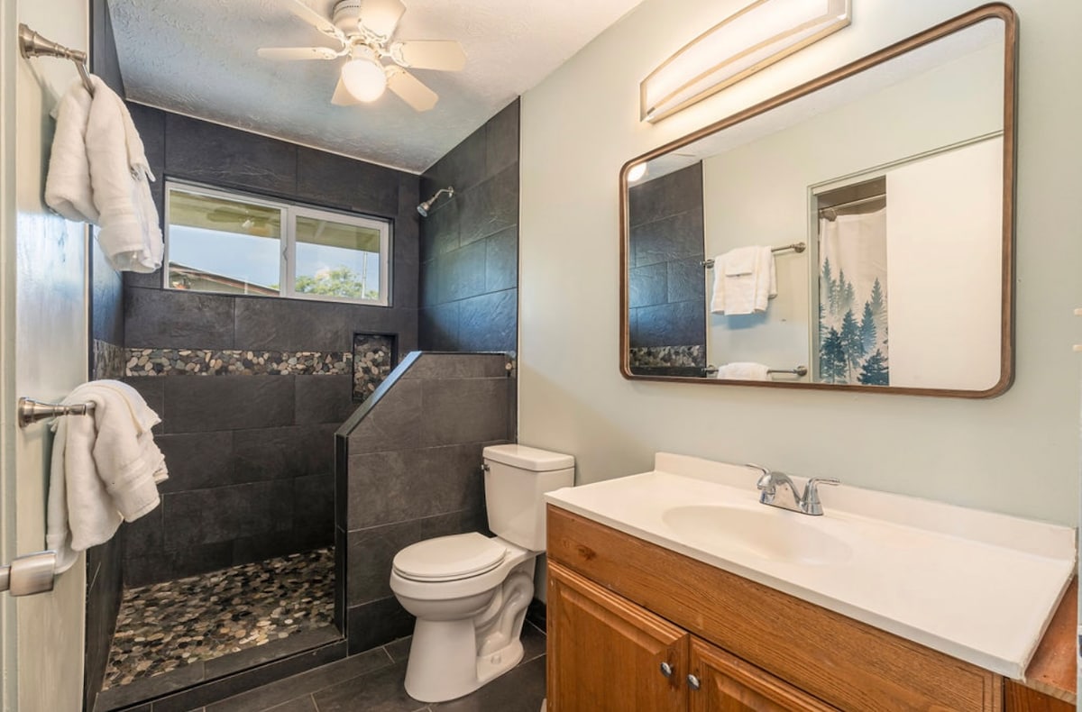 A contemporary bathroom features a dark tiled shower with a unique stone floor. A large mirror and light fixture are mounted above a wooden vanity, where toiletries may be placed. Two white towels hang on a bar, and a window allows natural light to enter.