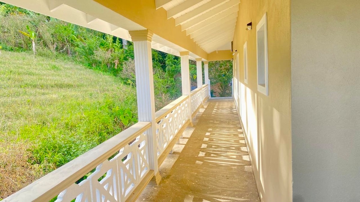 A spacious balcony is framed by white railings, overlooking lush greenery. Natural light casts soft shadows on the floor as the area connects to the upper-level apartment. The open space invites fresh air, providing a serene view of the surrounding landscape.