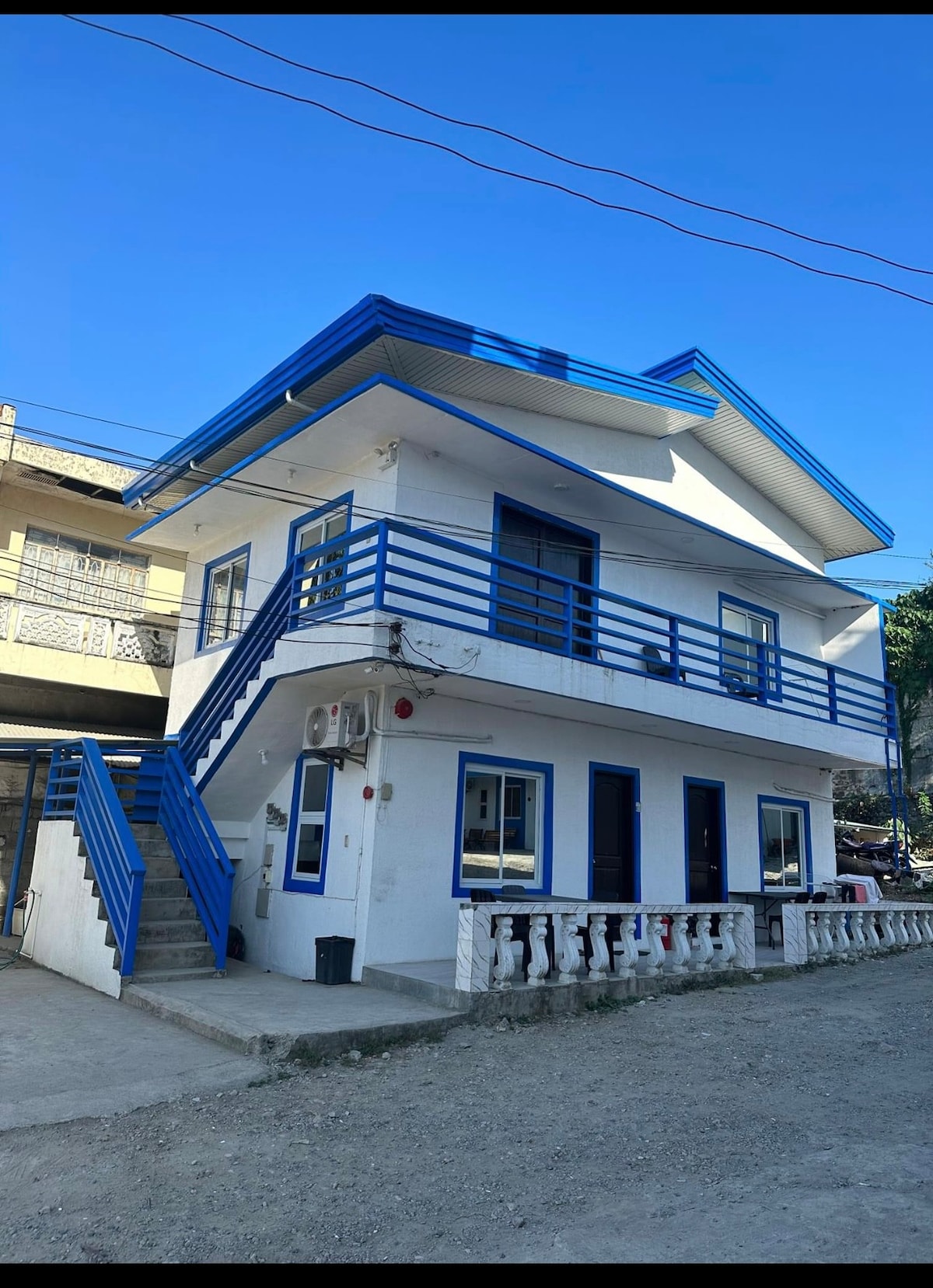 A two-story building showcases a bright blue and white exterior, highlighting a spacious balcony with blue railings. The structure is situated on a gravel pathway, featuring multiple windows that allow natural light to fill the interior.