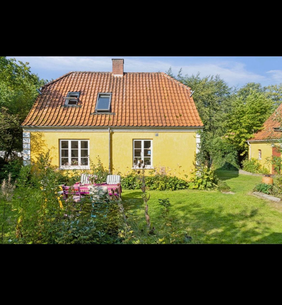 A charming yellow house features a red-tiled roof and multiple windows. A lush garden surrounds the home, showcasing green foliage and flowering plants. A patio area is visible with pink chairs set on the grass, inviting relaxation in a peaceful outdoor setting.