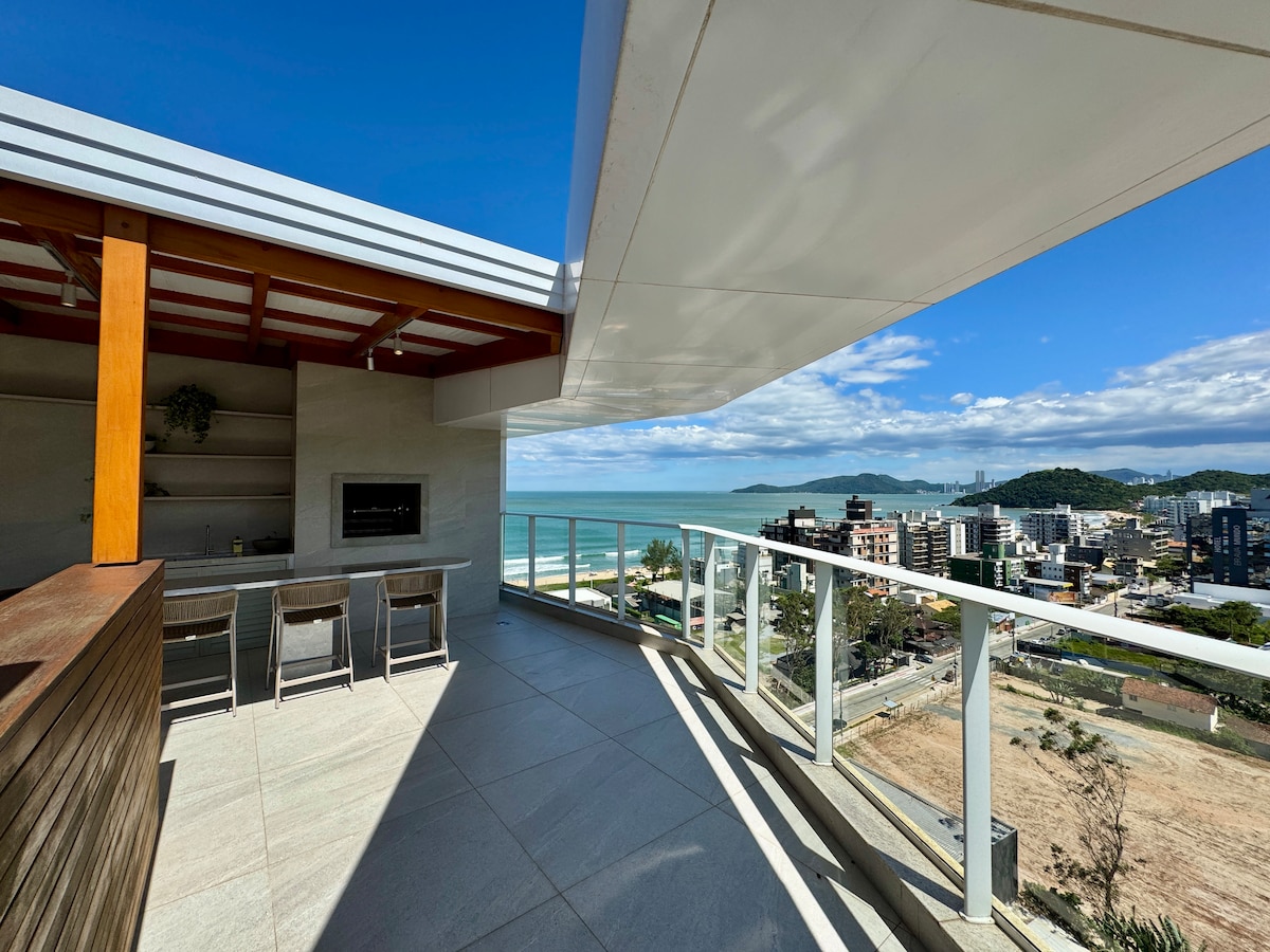 An expansive outdoor terrace offers stunning views of the ocean and surrounding landscape. The space features a built-in grill area with seating, complemented by a modern aesthetic. Large glass railings enhance the view and create an open atmosphere.