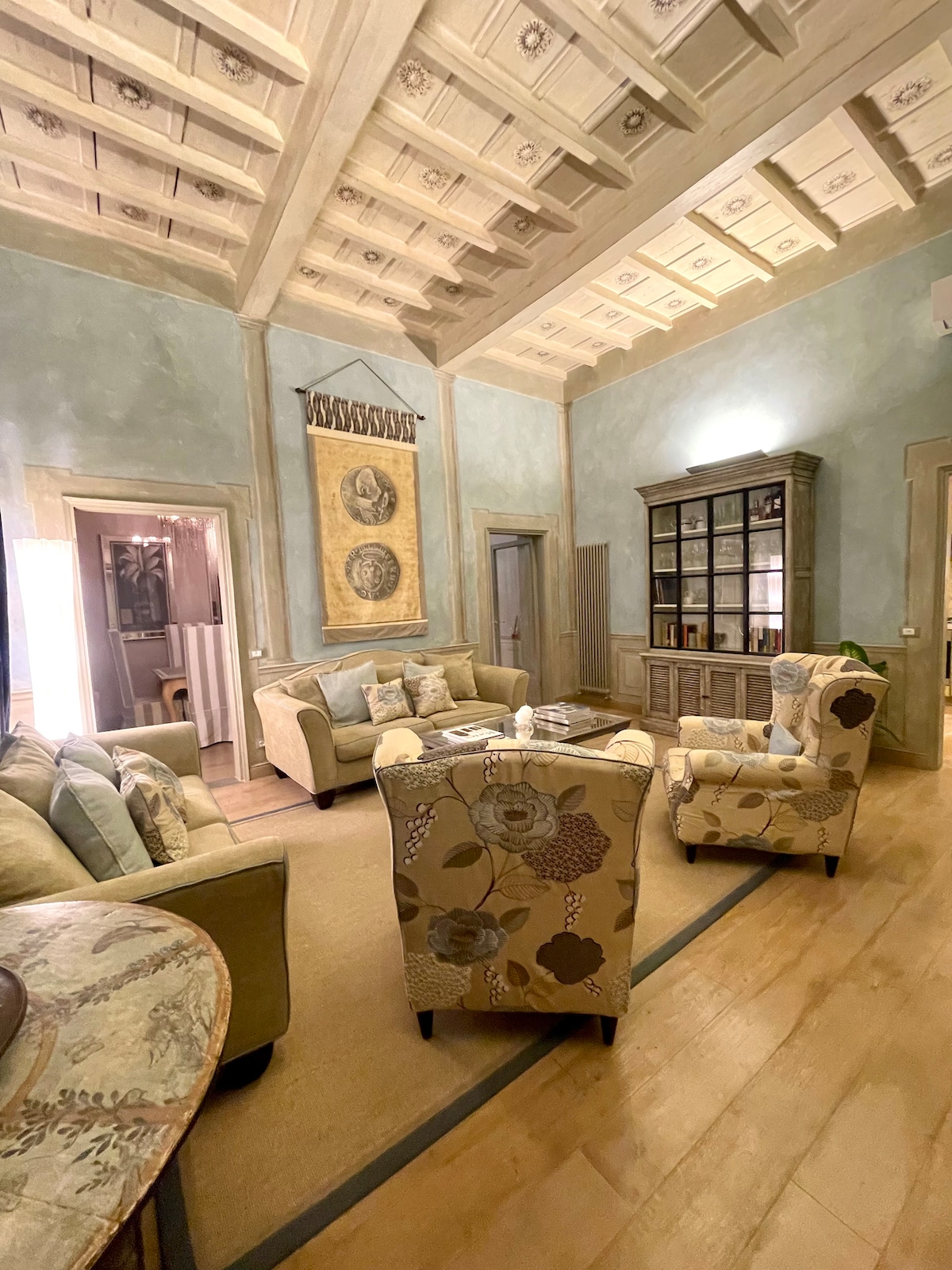 A living area features high ceilings adorned with decorative woodwork. Comfortable seating arrangements include two patterned armchairs and a spacious sofa. A wooden cabinet is centrally placed, showcasing an inviting collection of items. Soft lighting contributes to a calm environment.