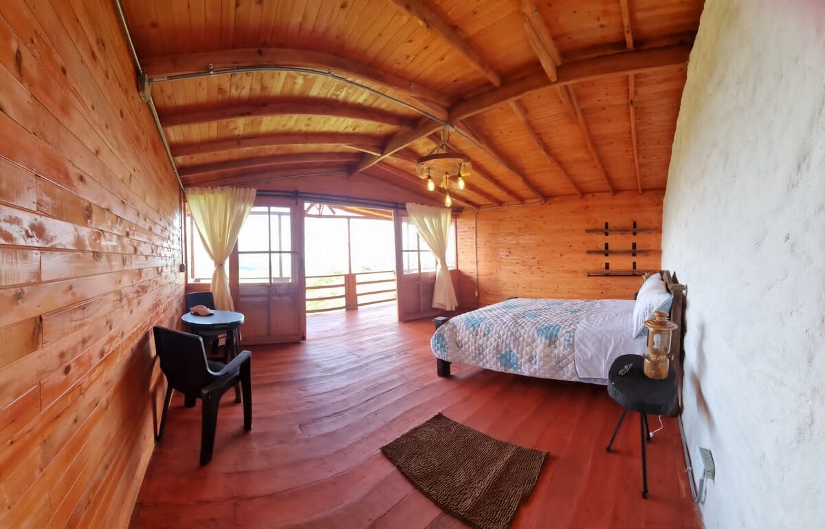 A spacious bedroom features wooden walls and ceiling, offering a warm ambiance. Large glass doors provide a scenic view and allow natural light to fill the room. A comfortable bed is complemented by a small table and a chair, with a woven mat on the floor.
