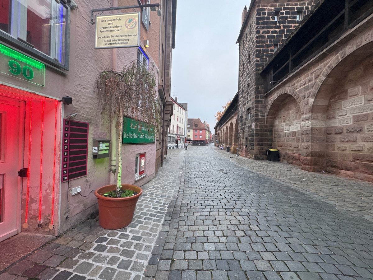 A quiet cobblestone street is bordered by historic buildings, showcasing a mix of textured stone and muted façades. A potted plant stands near the entrance of a building, while subtle red lighting adds interest to the scene. The atmosphere conveys a sense of urban charm.