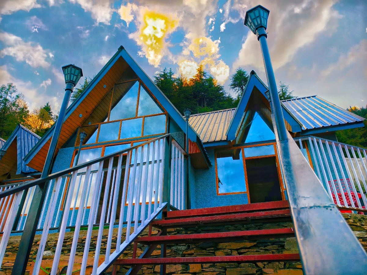 A charming cottage structure is depicted from a low angle, showcasing a sloped roof and large glass windows reflecting the sky. Two walkway lights frame the entrance, while a set of steps leads to the welcoming porch, surrounded by green trees.