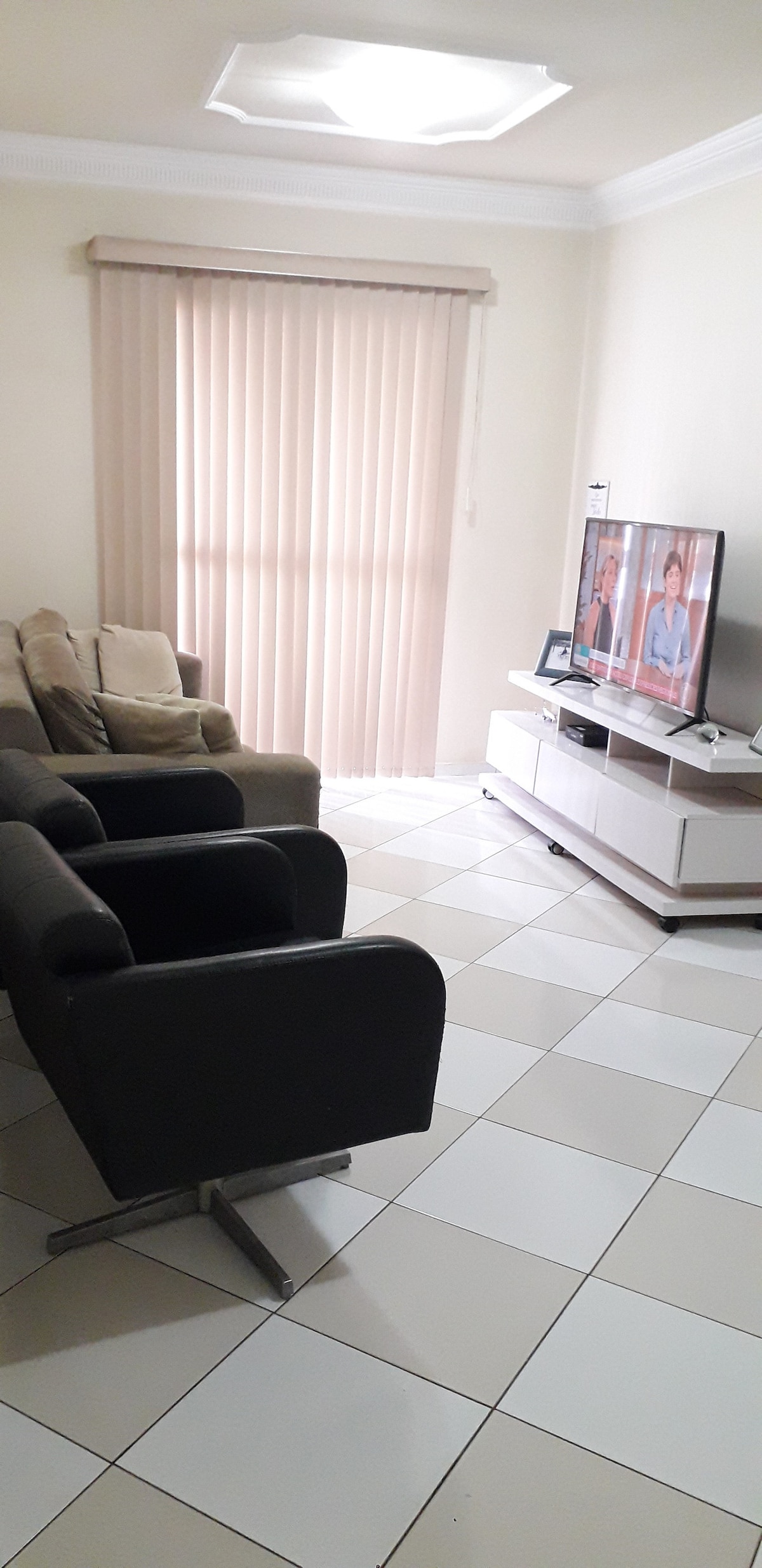 A living room features a set of two armchairs and a sofa, arranged around a modern television stand. Light-colored walls and flooring create a bright atmosphere. Vertical blinds cover a window, allowing natural light to filter into the space.