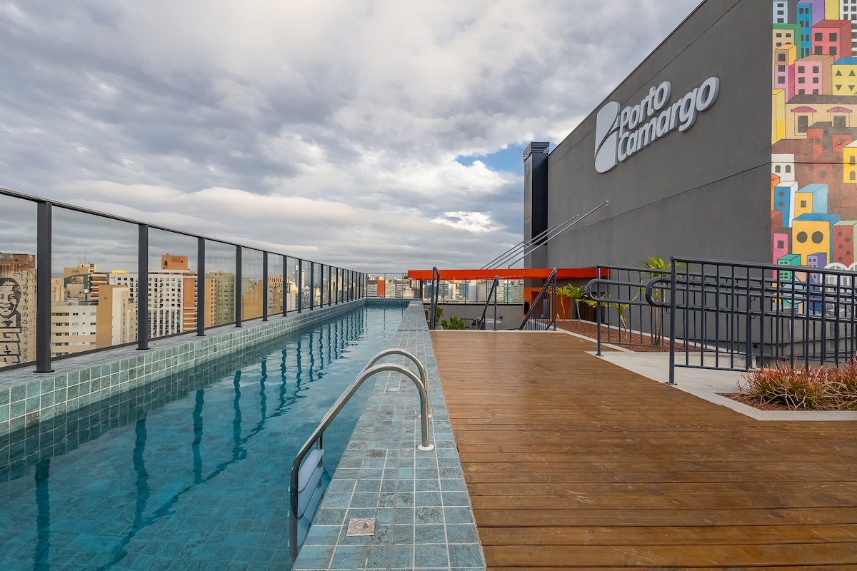 The rooftop area features a sleek swimming pool bordered by modern tiles and wooden decking. A vibrant mural decorates the adjacent building, adding a pop of color to the urban skyline. Lounge chairs are placed nearby, offering a space to relax and enjoy the view.