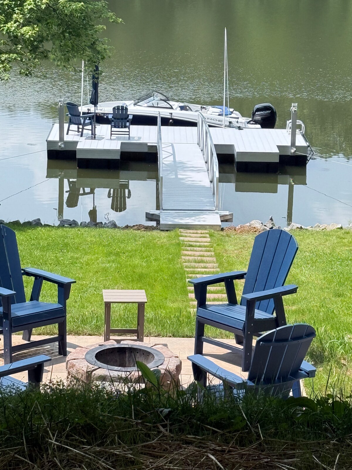 Two blue adirondack chairs face a small fire pit, positioned on a grassy area leading to a dock over still water. The dock holds a boat, and a pathway of stone pavers guides through the grass to the water's edge, surrounded by trees.
