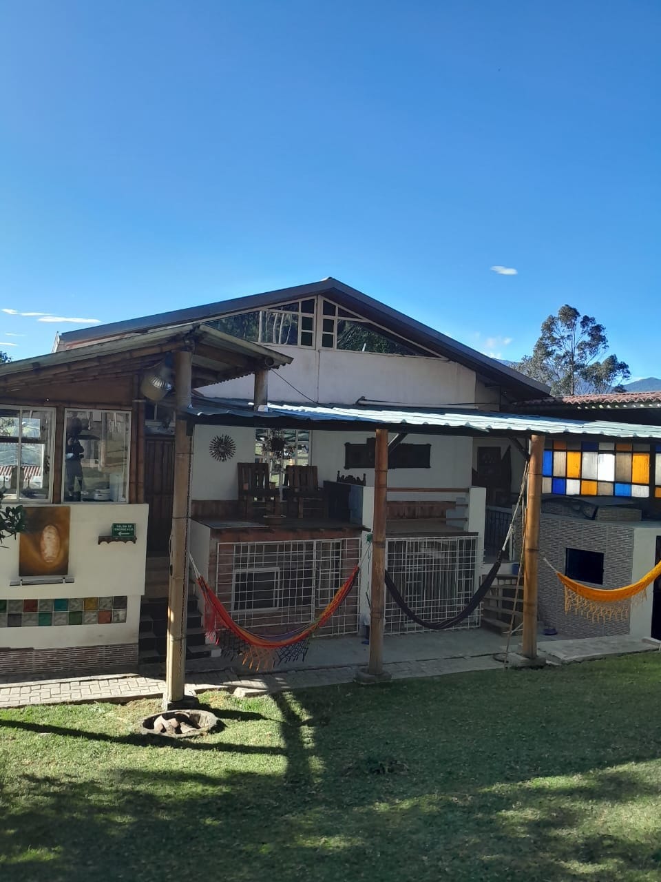 A charming house is framed by a bright blue sky, featuring large windows and a wooden terrace. Colorful hammocks are strung beneath the roof, inviting relaxation. Lush greenery surrounds the property, with a patio area that highlights the connection to nature.
