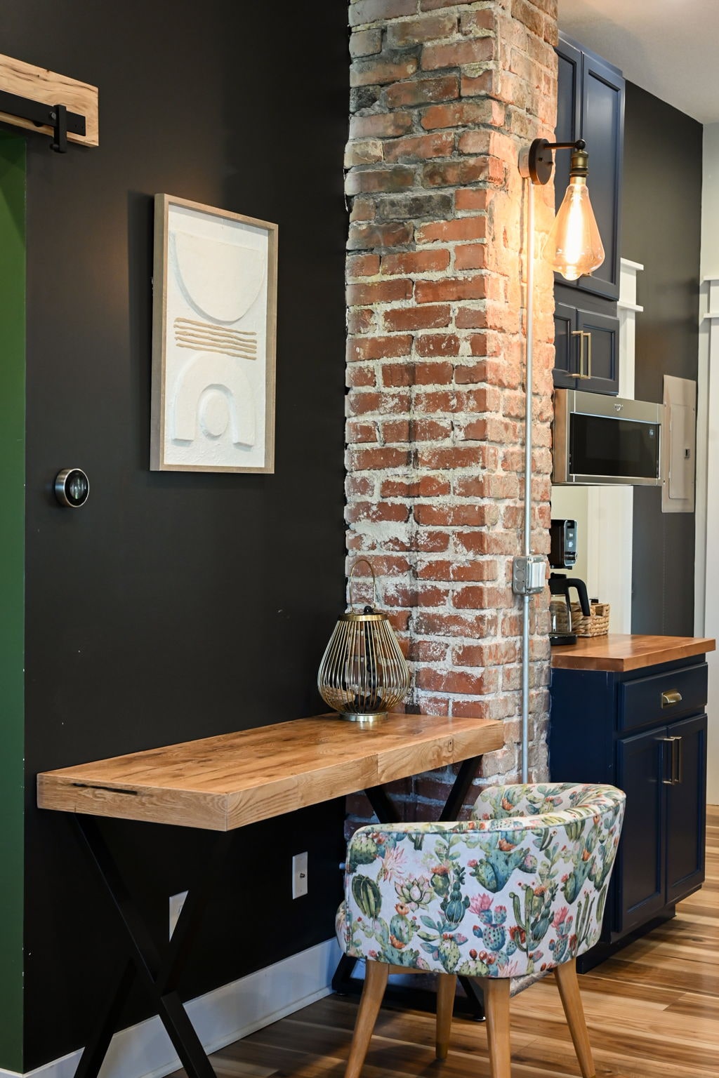 A small workspace is presented alongside a brick accent wall. A light fixture hangs above a wooden table, while a patterned chair adds a touch of color. Modern cabinetry and kitchen appliances are visible in the background, contributing to a functional and stylish environment.