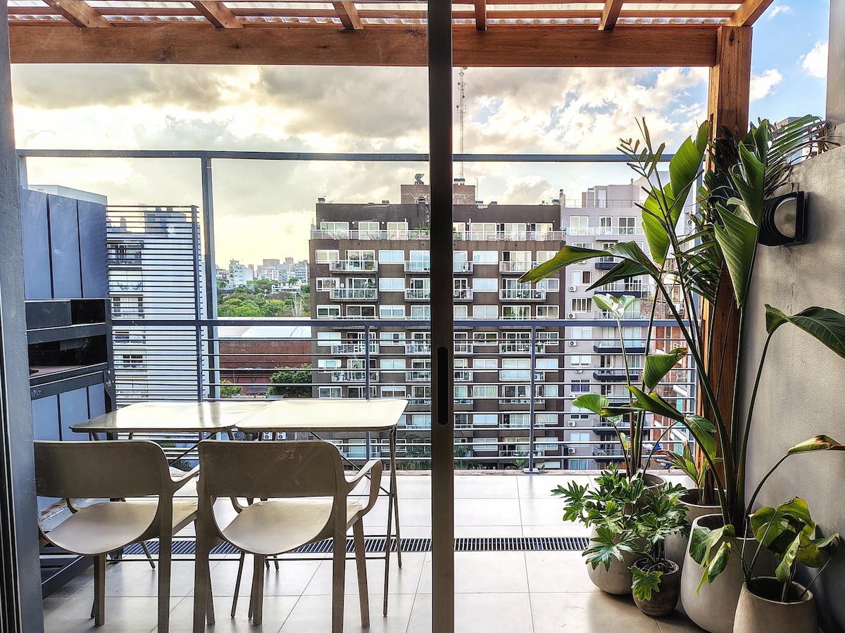 A private terrace features a light table and chairs, surrounded by various potted plants. The view offers a glimpse of the city skyline and nearby buildings, with a wooden pergola overhead allowing for shade against the sun.