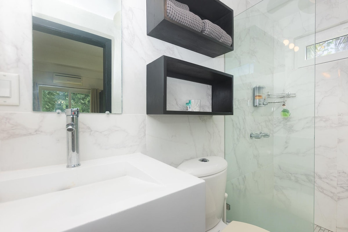 A modern bathroom is displayed, featuring a sleek white sink and a wall-mounted mirror. Black shelves hold neatly arranged towels, while a frosted glass shower enclosure is visible. Soft, natural light filters in through a small window.