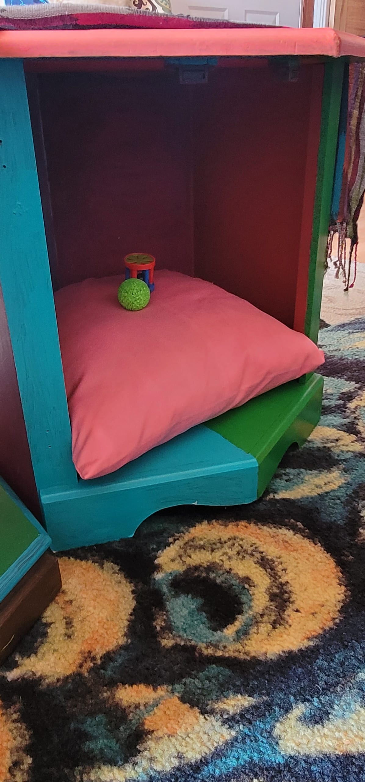 A colorful storage unit featuring a vibrant blue and red design houses a soft pink cushion. A small green toy is positioned on the cushion, adding a playful touch to the inviting space. The surrounding area is adorned with a patterned area rug.