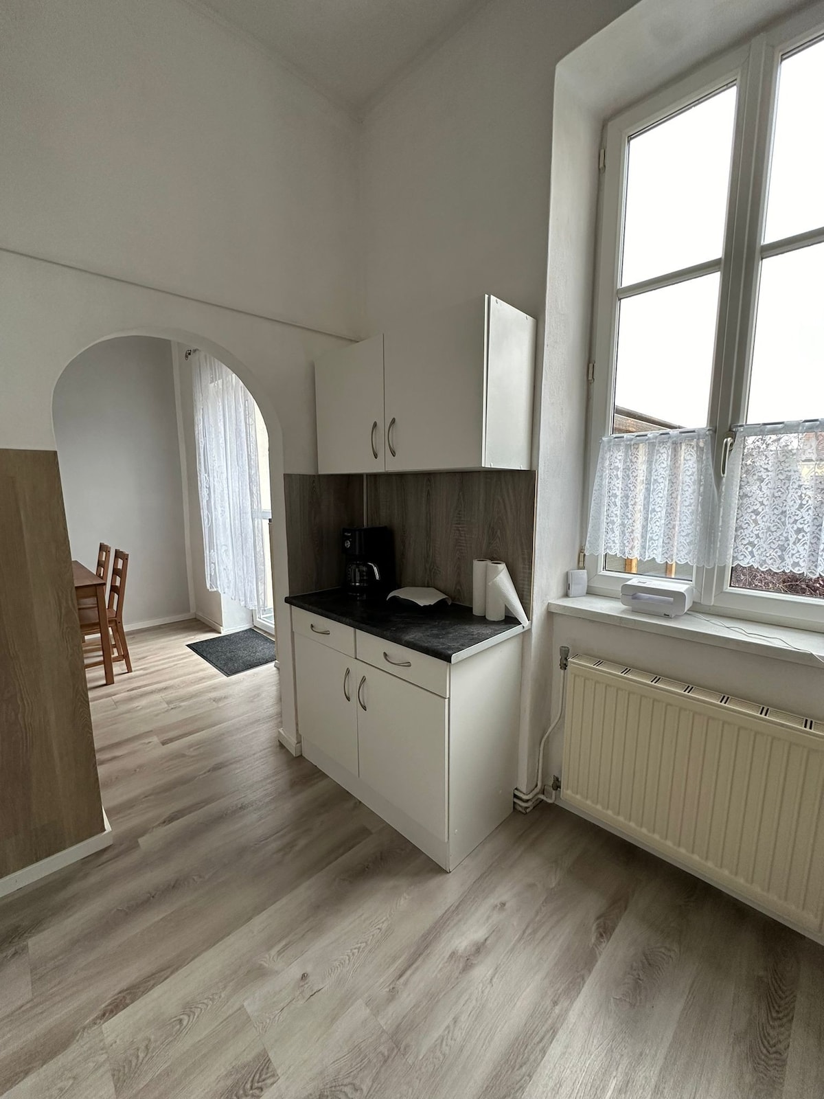 A compact kitchenette is shown with white cabinetry and a dark countertop. A coffee maker sits on the counter, and natural light enters through a window adorned with lace curtains. An archway leads to a nearby dining area, visible in the background.