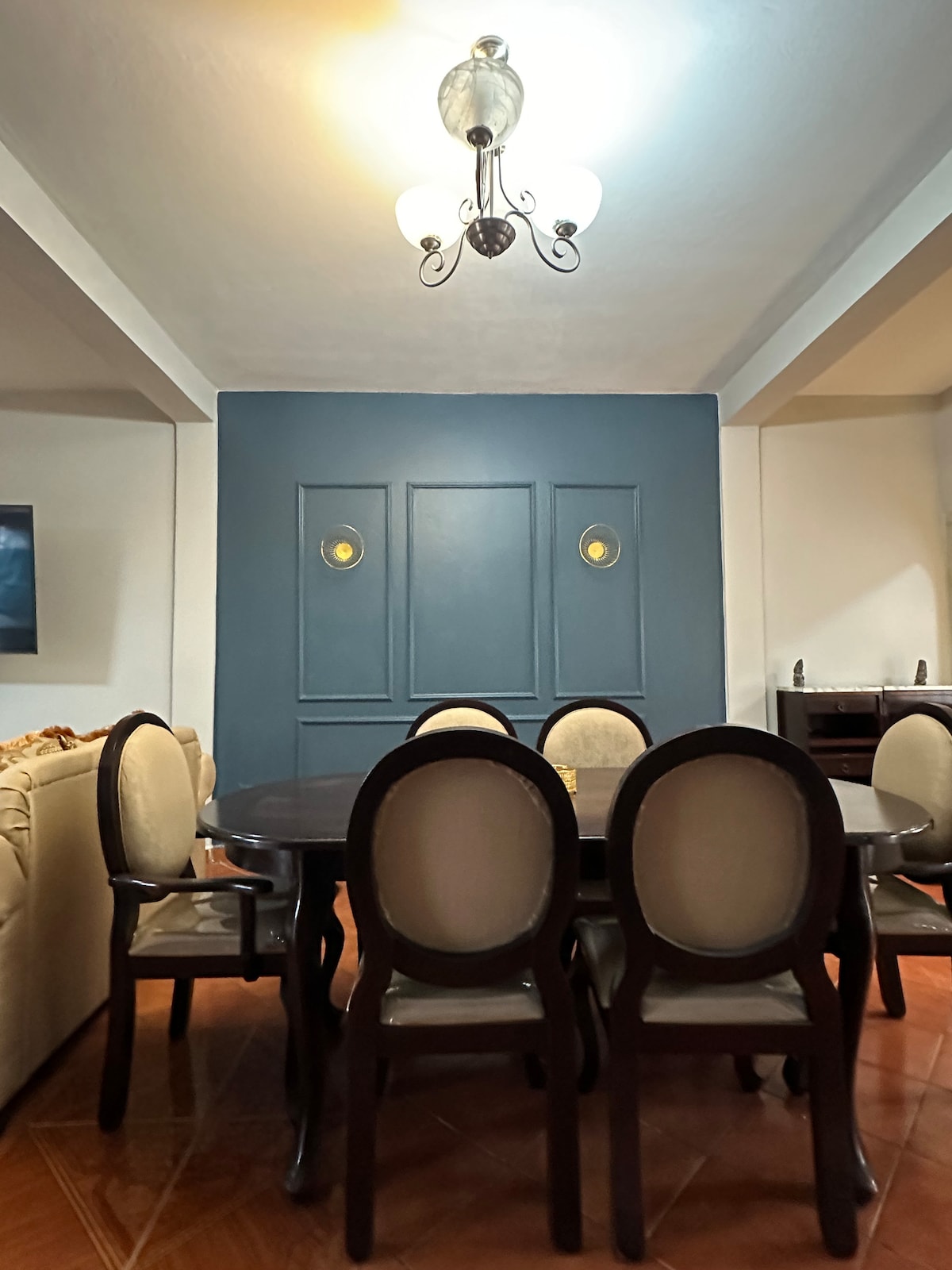 A dining area features a round table surrounded by six upholstered chairs. A stylish chandelier hangs from the ceiling, providing soft overhead lighting. The backdrop includes a blue accent wall with two circular decorative elements, adding depth to the space.