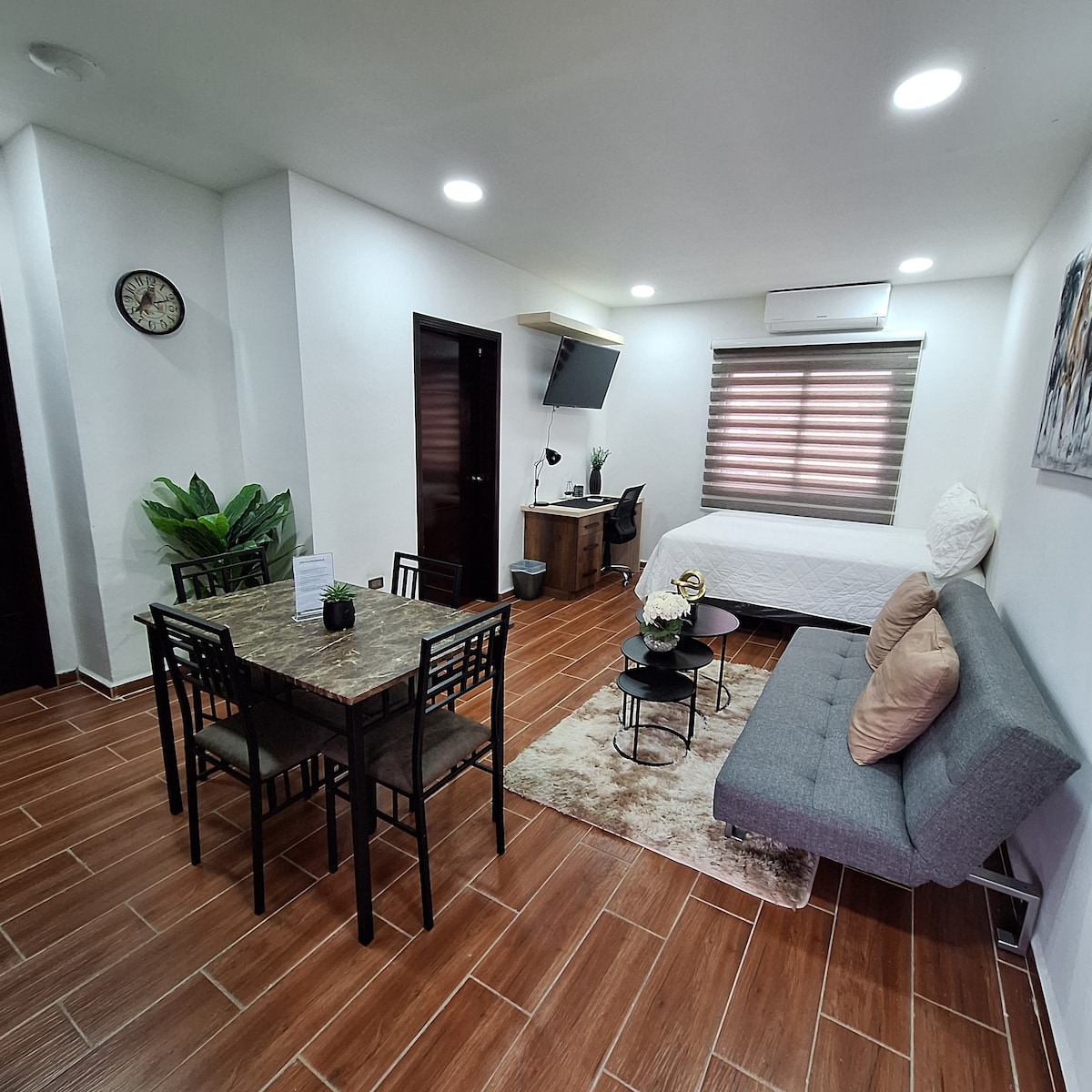 An inviting living space features a gray sofa and a dining table with six chairs. A bed is situated in the background, complemented by a desk and chair. Natural light filters through window shades, and a decorative rug lies on the floor.