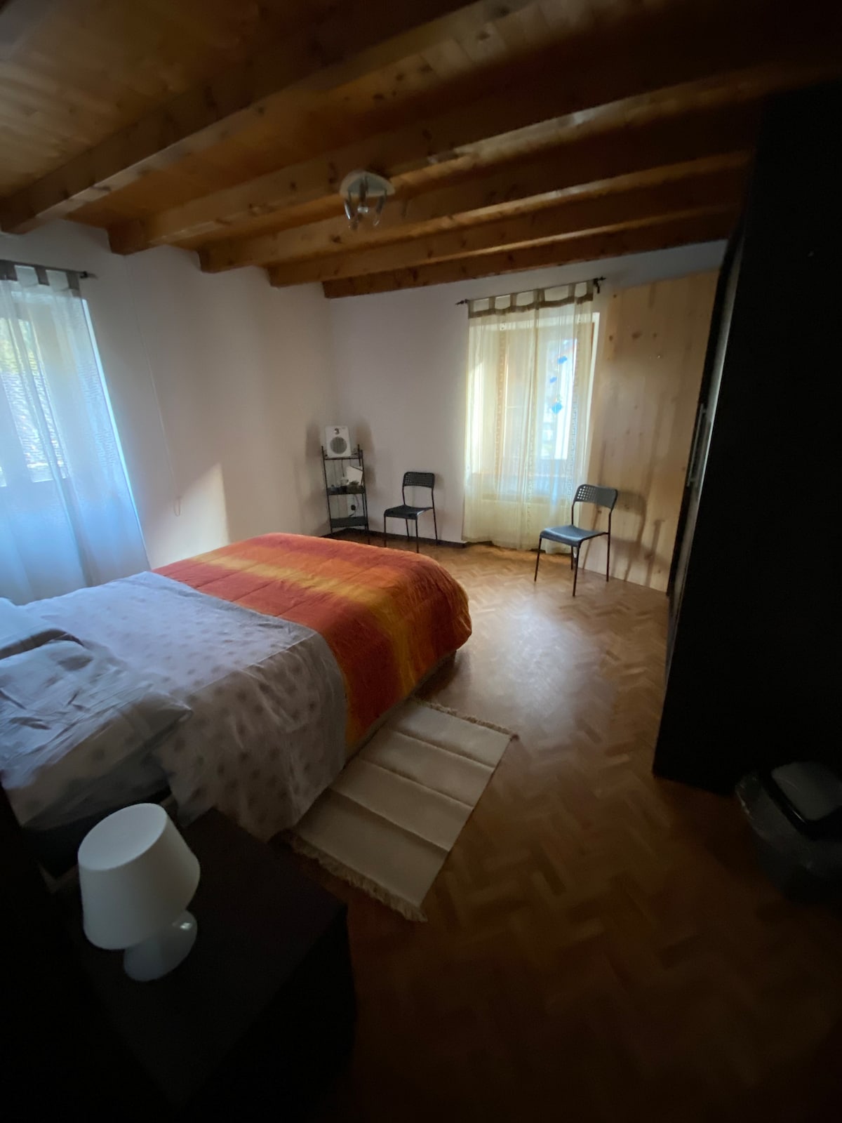 A bedroom features a warm-toned bedspread draped over a spacious bed. Two chairs are positioned beside the bed, and light filters through sheer curtains at the windows. Wooden beams draw attention to the ceiling, while a nightstand holds a lamp, enhancing the room's comfort.