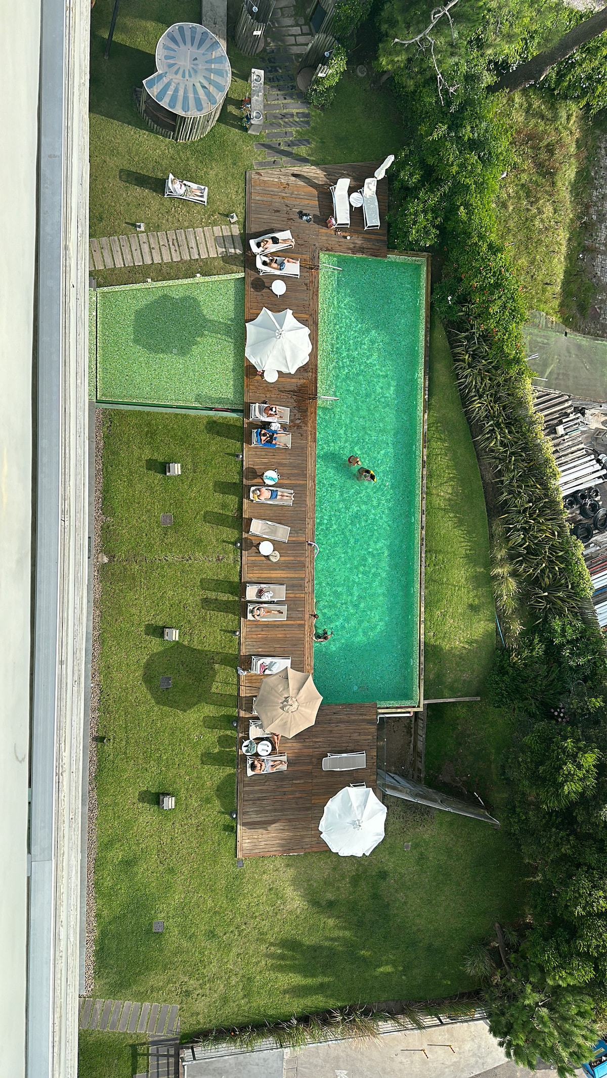 An aerial view showcases a rectangular swimming pool surrounded by a wooden deck, sun loungers, and umbrellas. Green grass lines the perimeter, with scattered lounge areas and a small gazebo visible nearby, offering a serene outdoor environment.