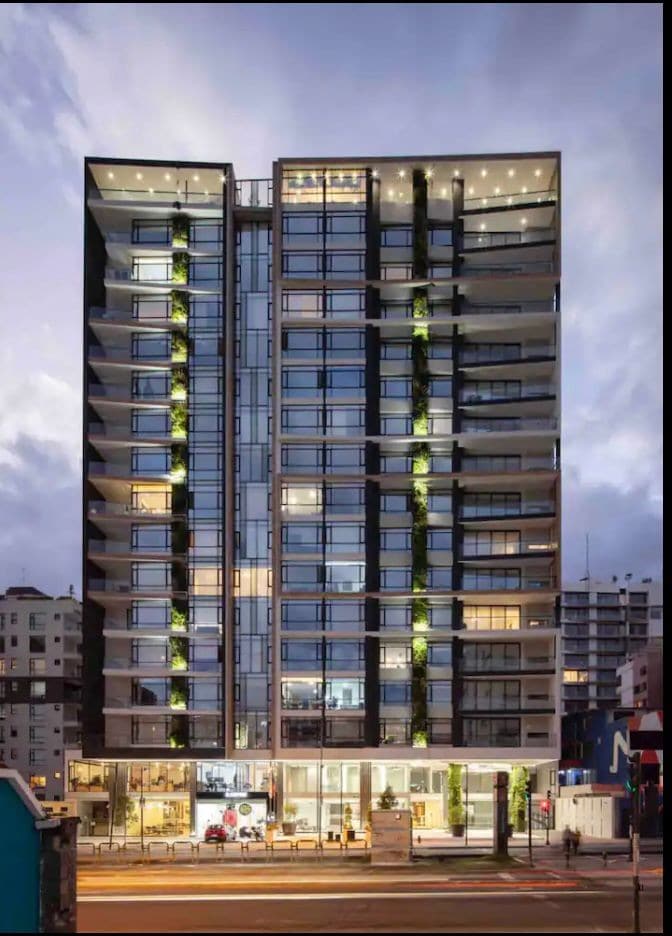 A modern high-rise building features a facade of large glass windows, providing a glimpse of the stylish interiors. The structure is adorned with greenery along the balconies and illuminated by lights, creating a sophisticated urban presence at dusk.