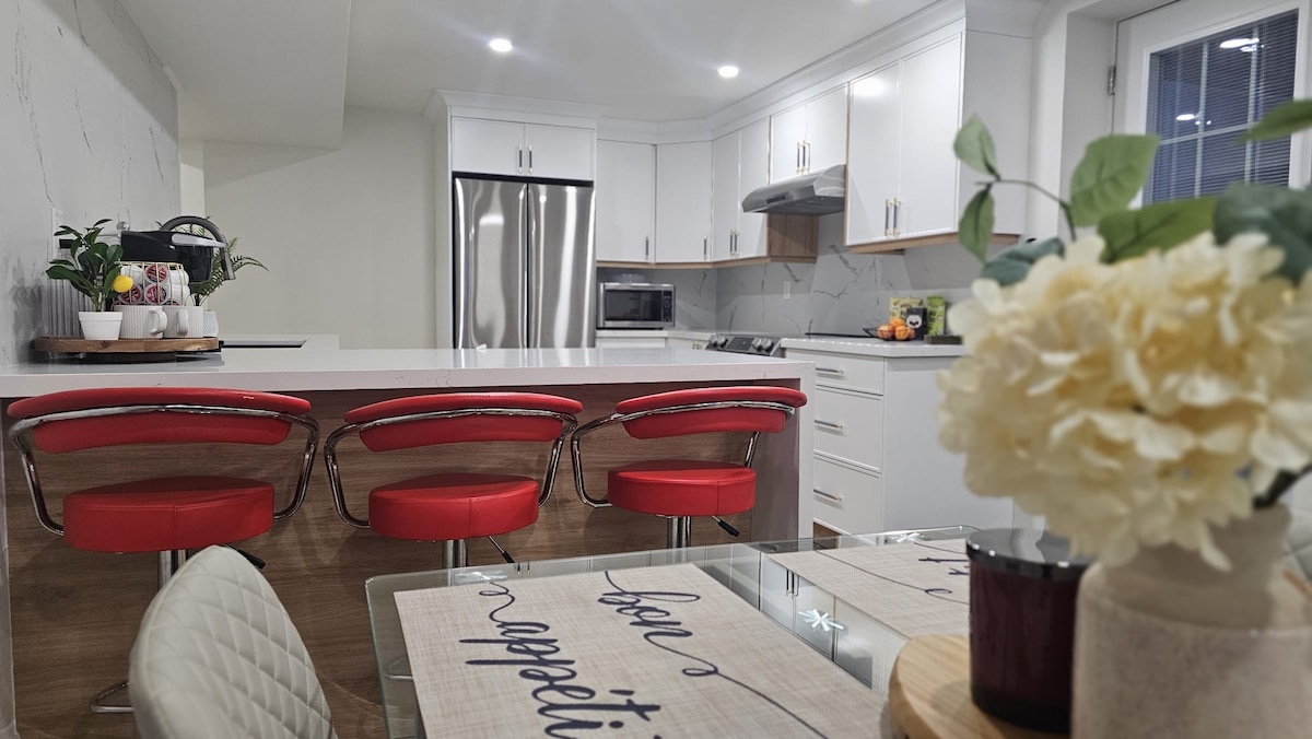 The kitchen showcases modern cabinetry and sleek countertops, featuring a spacious island with three red stools. Stainless steel appliances, including a refrigerator and microwave, complement the contemporary design. A dining table adorned with a neutral placemat and a vase of flowers adds a welcoming touch.