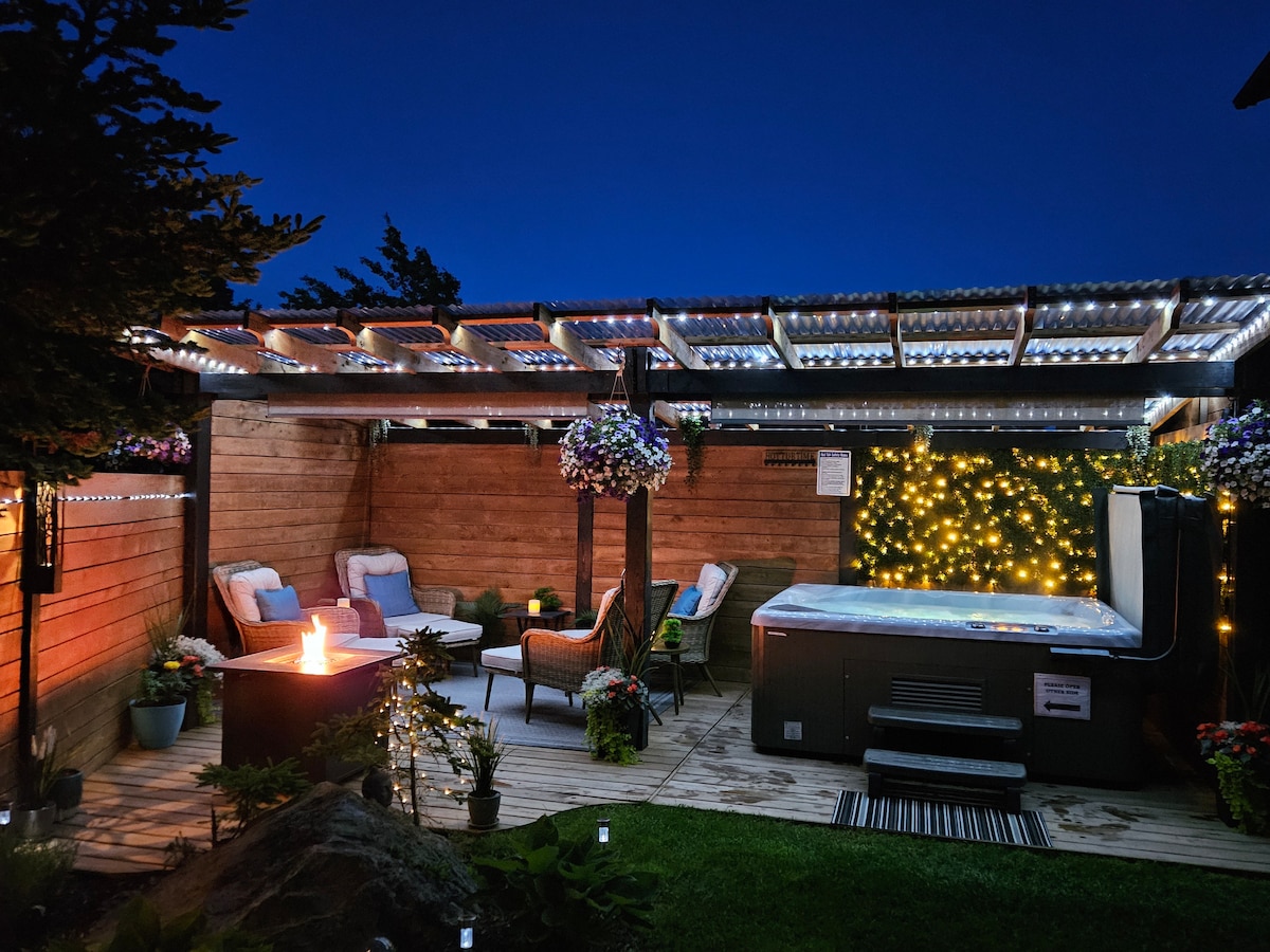 A serene outdoor space is illuminated by soft lights against a night sky. A covered pergola shades a relaxing hot tub, while a cozy seating area with a firepit invites conversation. Lush plants and colorful hanging flowers enhance the peaceful ambiance.