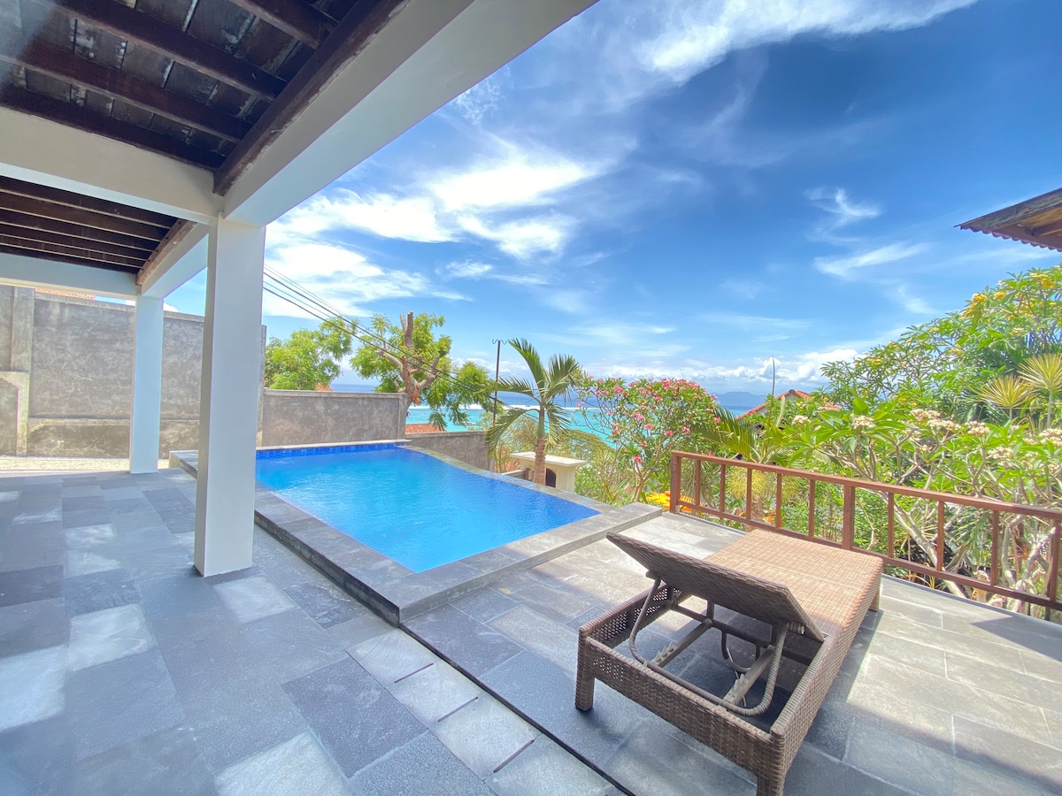 A private pool is set against a backdrop of lush greenery and tropical views. Sun loungers are positioned next to the pool on a stone patio, providing a comfortable space to relax. The scene is framed by an open area that enhances the connection to the outdoors.