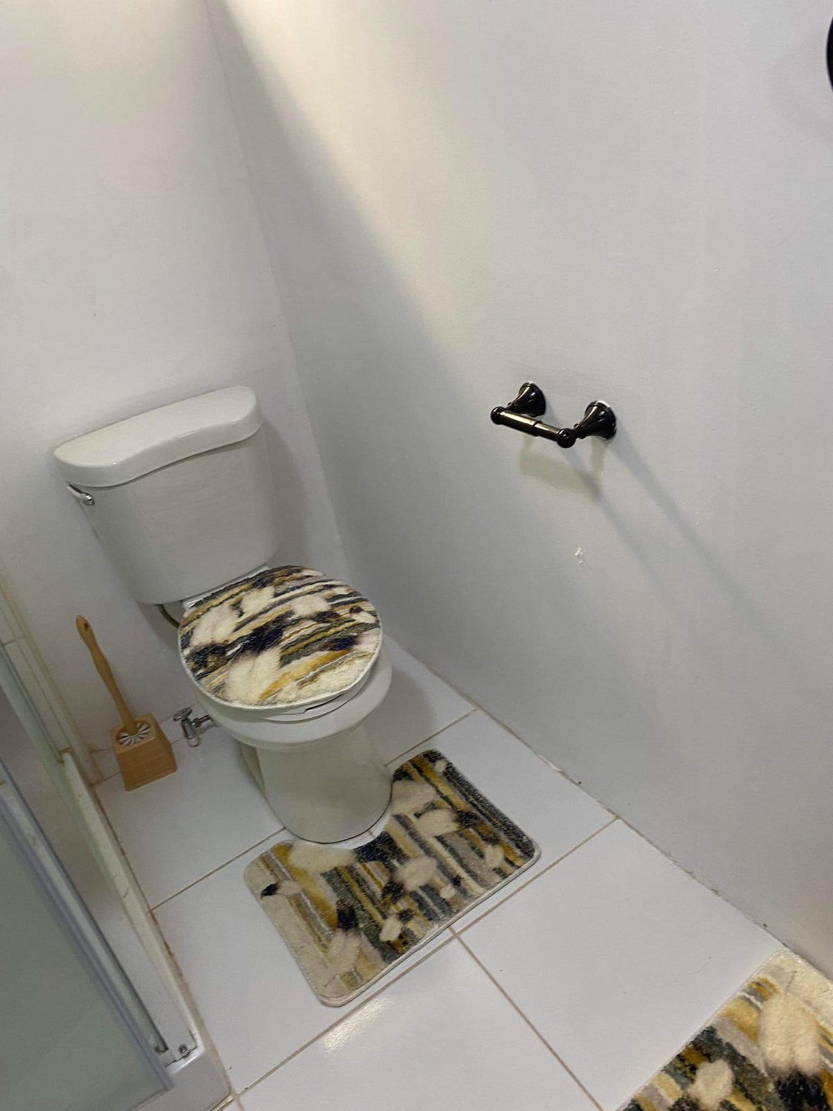 A clean bathroom is depicted, featuring a modern toilet with a decorative cover. Soft rugs are placed on the tiled floor, and a wall-mounted towel bar is visible nearby. A wooden toilet brush holder adds to the organized space.