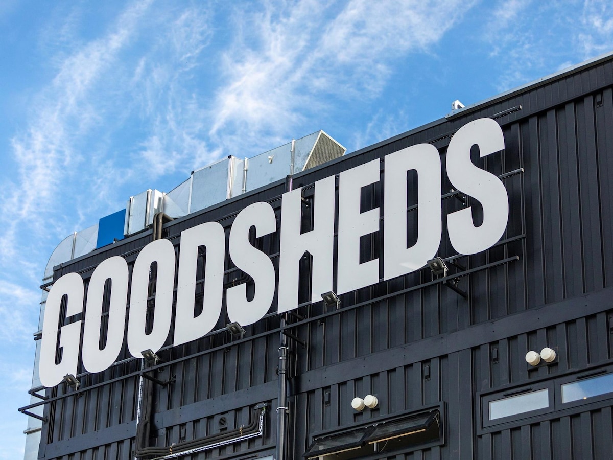 The exterior of the Goodsheds building is showcased, featuring a large, bold sign displaying the name 'GOODSHEEDS' in uppercase letters. The building's facade is sleek and modern, contrasted against a bright blue sky with scattered clouds.