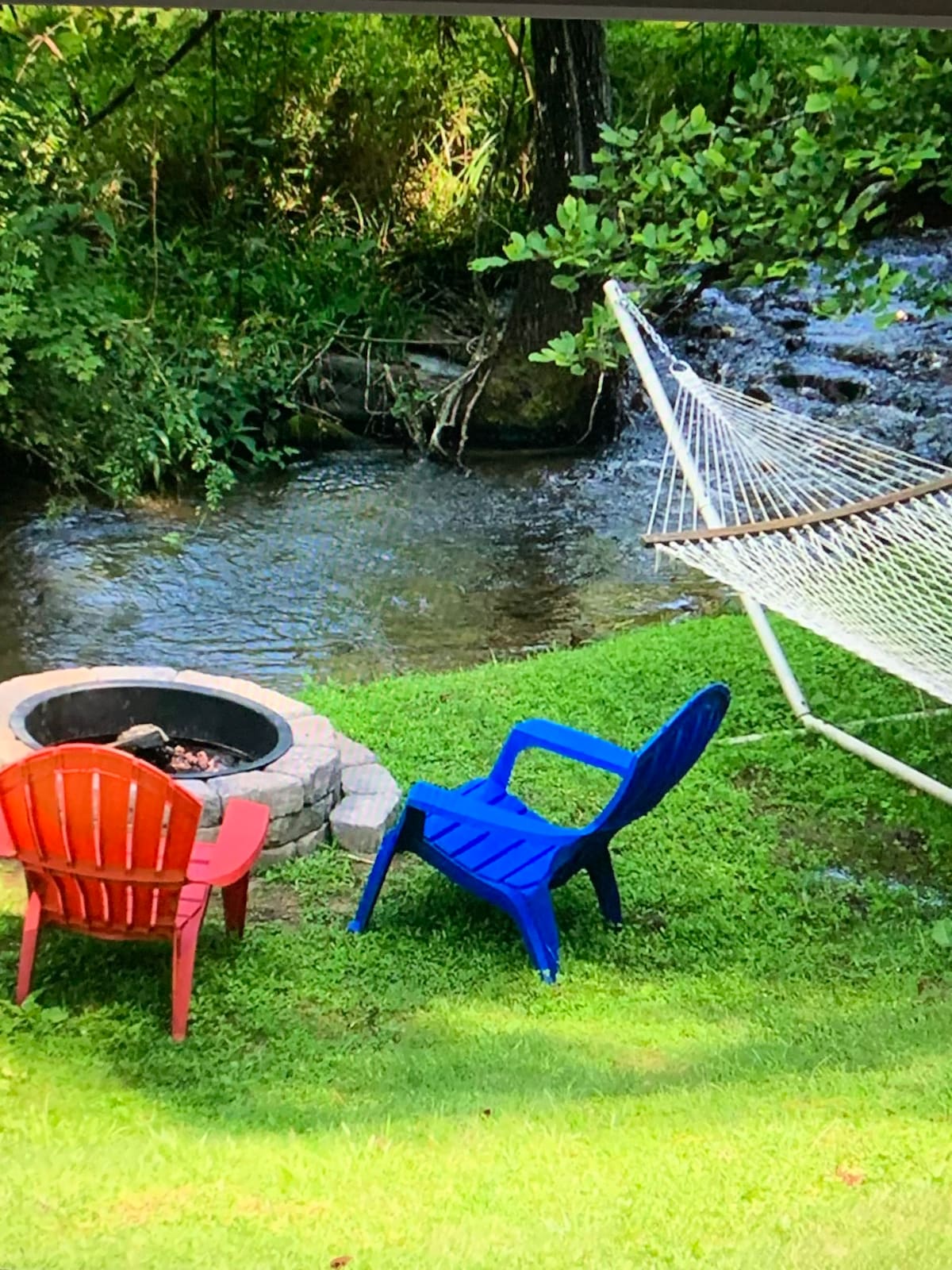 A serene outdoor space features a fire pit surrounded by two adirondack chairs in red and blue. Nearby, a hammock is strung between trees, gently swaying. A creek flows quietly in the background, enhancing the peaceful ambiance of the lush green landscape.