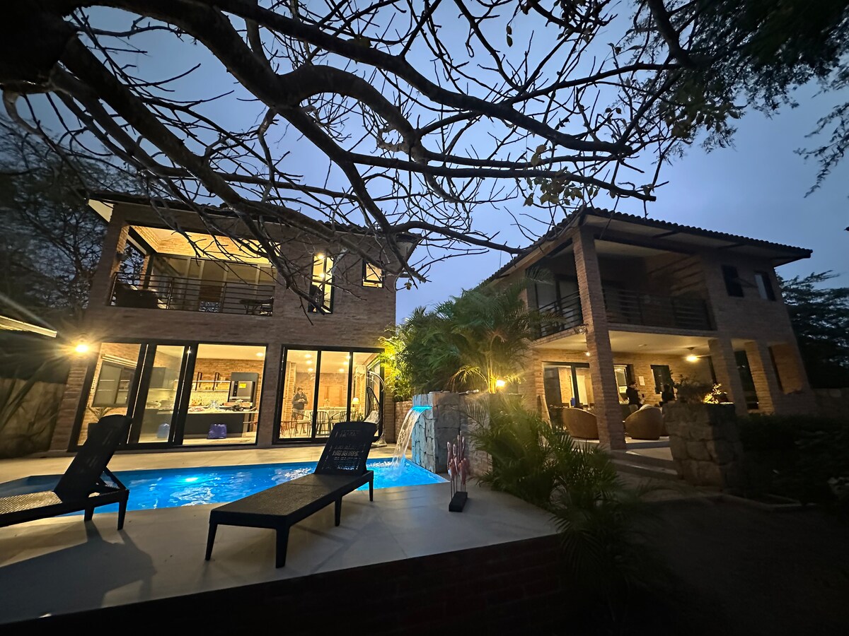 Two modern houses are set within a landscaped area, illuminated softly at dusk. A sparkling pool is positioned in the foreground, bordered by lounge chairs. Expansive glass doors reveal cozy interiors. Lush plants provide greenery around the amenities.
