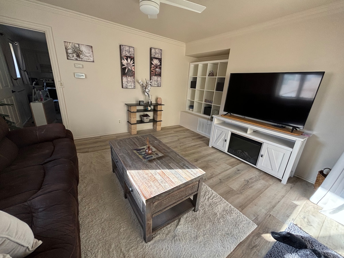 A well-appointed living area features a dark leather sofa paired with a rustic coffee table at the center. A large television is mounted on a white stand, and decorative shelves are displayed against the wall, complemented by framed art pieces.